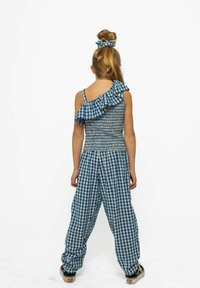 Lány hátat fordítva áll, kék és fehér kockás, egy vállon viselt felsőt és hozzá illő laza nadrágot visel, haja kontyba van fogva egy hajgumi segítségével.