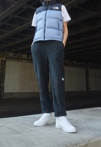 Niebiesko-czarna pikowana kamizelka na białym t-shircie, zestawiona z ciemnymi dresami z czarnymi łatami na kolanach oraz białymi trampkami na betonowej nawierzchni.