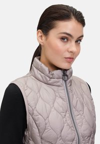 Licht taupe geborduurd mouwloos vest met hoge kraag, voorritssluiting en contrasterende zwarte armsgaten. Zachte textuur met subtiele glans.