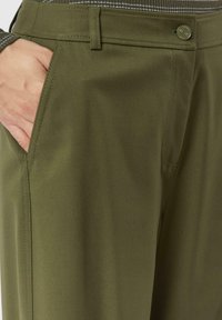Spodnie w kolorze oliwkowej zieleni o gładkiej fakturze, krój dopasowany, z przednimi kieszeniami i detalem kontrastującego przeszycia. Z przodu zapięcie na guziki.