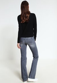 Kobieta z długimi brązowymi włosami, ubrana w czarną bluzkę z długim rękawem, wyblakłe niebieskie dżinsy typu bootcut i białe trampki, stojąca tyłem na jednolitym tle.