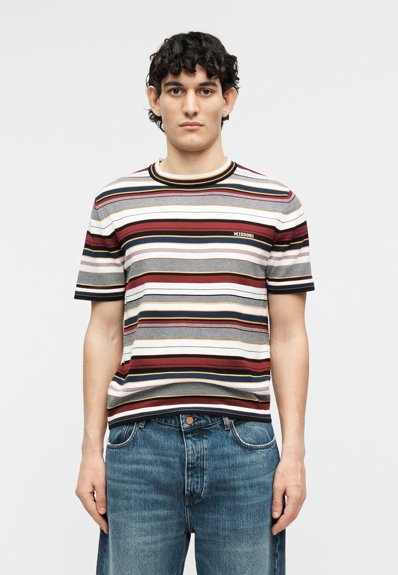 Man wearing a short-sleeve knit shirt with horizontal stripes in red, white, black, and yellow paired with blue jeans, standing facing forward.