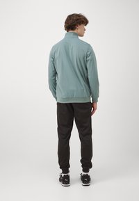 Lys grøn pullover med høj krave, parret med sorte tapered bukser og sorte sportssko, set fra ryggen.