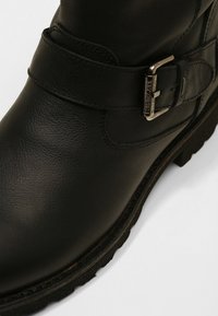 Svart läderankelstövel med texturerad yta, prydd med ett brett spännband och en robust sula. Närbild som framhäver designens detaljer.