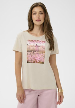 Žena v béžovom tričku s potlačou dievčaťa v ružových šatách stojaceho pri zábradlí obklopenom ružovými kvetmi.