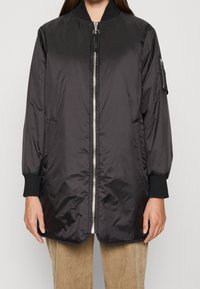 Černá, lehká bomber bunda s předním zipem, žebrovanými manžetami a kapsou na prsou. Vyznačuje se hladkým, lesklým povrchem a střední délkou.