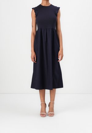 Mujer con vestido midi azul marino sin mangas con detalles de volantes en los hombros y tacones beige abiertos con correa en el tobillo, de pie frente a un fondo blanco.
