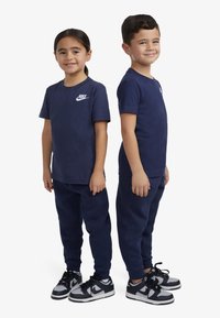 T-shirt in cotone blu navy con logo Nike bianco, abbinato a pantaloni della tuta blu navy e sneakers in bianco e nero. Bambini in piedi uno accanto all'altro.