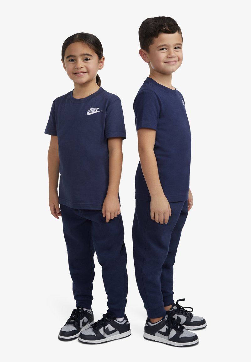 T-shirt in cotone blu navy con logo Nike bianco, abbinato a pantaloni della tuta blu navy e sneakers in bianco e nero. Bambini in piedi uno accanto all'altro.