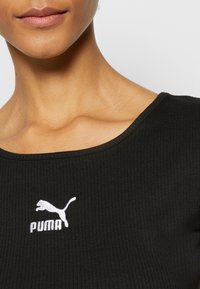 Gros plan sur une personne portant un haut côtelé noir avec un logo blanc Puma sur la poitrine et un col arrondi.