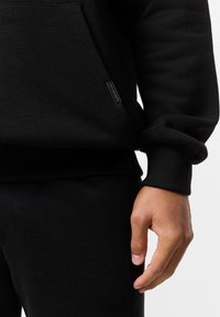 Zwarte katoenen hoodie met een kangoeroezak, geribbelde manchetten en zoom. Labeldetail aan de zijkant voegt een subtiel accent toe. Gecombineerd met bijpassende zwarte broek.