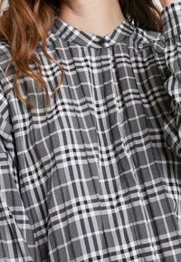 Svart och vit rutmönstrad blus med ståkrage, knappstängning och plisserade detaljer. Mjuk tyg med varierande små och stora rutor.