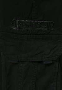 Svart klädsel med ett texturerat tyg som har en framträdande, broderad "ALPHA"-etikett. Inkluderar fickor med klaffstängningar och förstärkta sömmar.