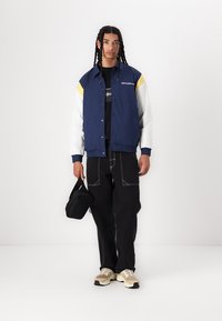 Veste de l'équipe bleu marine avec des accents blancs et jaunes, associée à un pantalon cargo noir et des baskets beiges, tenant un petit sac noir.