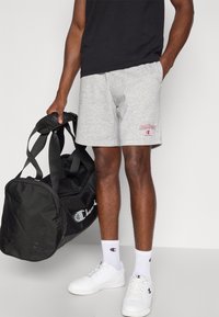 Γκρι βαμβακερά σορτς με κόκκινο λογότυπο «Champion», συνδυασμένα με λευκά αθλητικά παπούτσια και κάλτσες, κρατώντας μια μαύρη τσάντα duffel με λευκό λογότυπο.