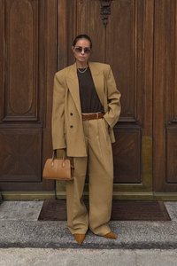 Woman wearing oversized tan suit, brown top, sunglasses, and pointed shoes, holding a tan handbag, standing in front of wooden doors.