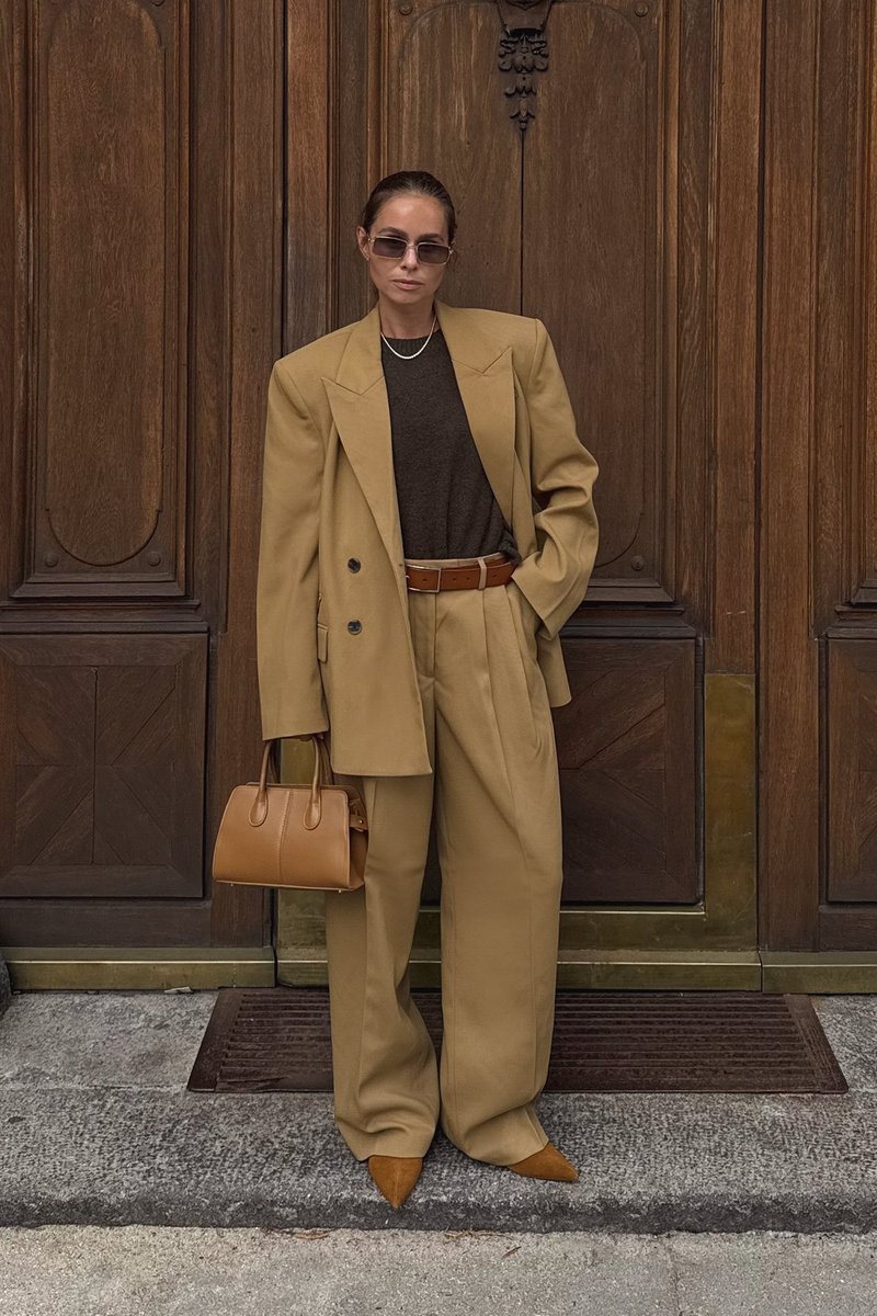 Woman wearing oversized tan suit, brown top, sunglasses, and pointed shoes, holding a tan handbag, standing in front of wooden doors.