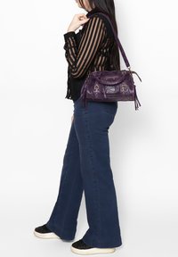 Paarse leren handtas met gouden hardware, franjes en een textuur oppervlak. Model draagt een zwart gestreepte top en donker uitlopende jeans.