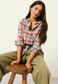 Plaid button-up shirt in red, blue, and cream, with rolled sleeves; paired with olive green drawstring pants. Wood stool with rustic texture.