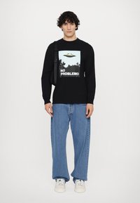 Black long-sleeve shirt with a graphic print featuring an alien spaceship and "NO PROBLEMO" text; worn with baggy light blue denim jeans.