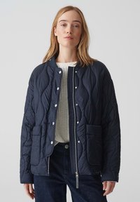 Giacca trapuntata blu navy con colletto alto, bottoni a pressione, due tasche frontali e chiusura con zip. Indossata sopra un maglione testurizzato color crema.