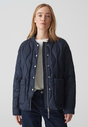 Navy quilted jacket with a stand collar, snap buttons, two front pockets, and a zip closure. Worn over a cream textured sweater.