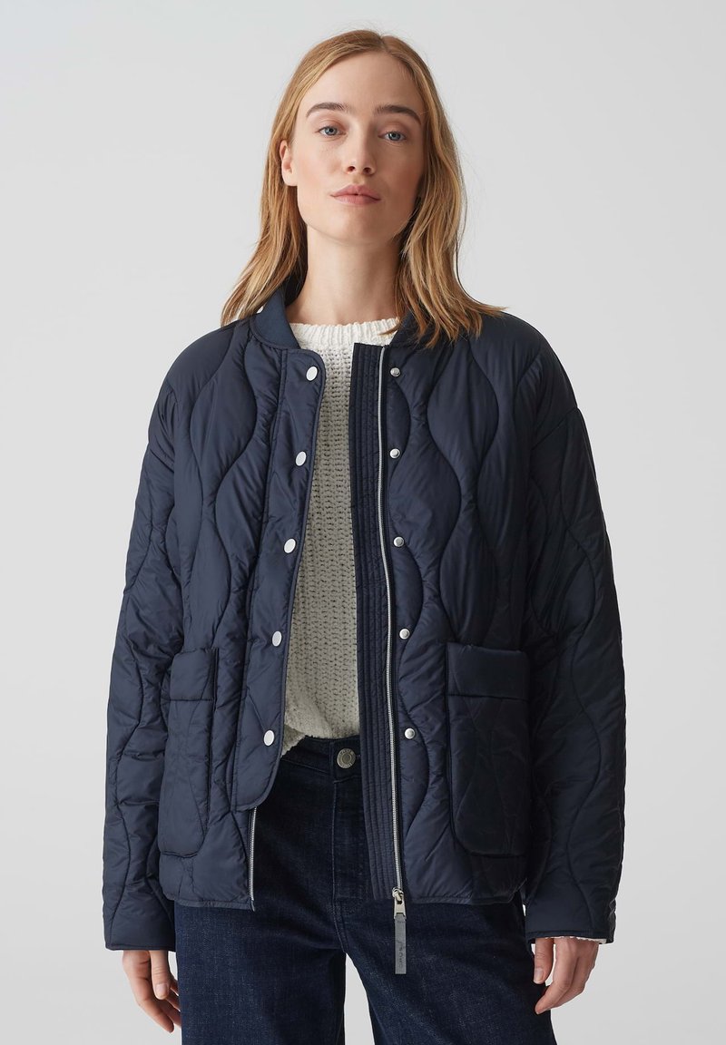 Giacca trapuntata blu navy con colletto alto, bottoni a pressione, due tasche frontali e chiusura con zip. Indossata sopra un maglione testurizzato color crema.