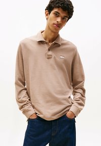 Langarm, hellbraunes Poloshirt aus strukturiertem Stoff, mit einem Kragen und einem kleinen Logo auf der linken Brust. Getragen mit dunklen Jeans.