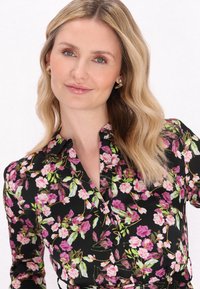 Sort blomsterkjole med pink og grønne blomster og en kravenakke. Modellen bærer guld blomsterøreringe og har bølget blond hår.