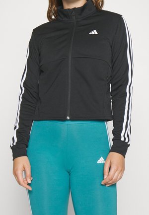 Femme portant une veste de sport noire zippée avec des bandes blanches sur les manches et un legging Adidas bleu sarcelle avec le logo sur la cuisse gauche.