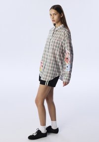 Giovane donna in piedi con camicia a quadri con toppe floreali sulle maniche, pantaloni shorts neri con strisce bianche, calze bianche e sneakers nere su sfondo bianco.