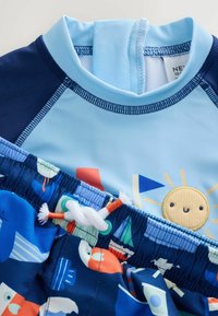 Colorato set da bagno per bambini con top azzurro chiaro decorato con un design solare e pantaloncini blu navy con motivi di onde e barche. Tessuto morbido ed elastico.