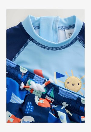 Krāsains bērnu peldkostīms, kas sastāv no gaiši zila augšdaļas ar saules dizainu un tumši zila apakšdaļas ar viļņu un laivu rakstu. Mīksts, elastīgs audums.