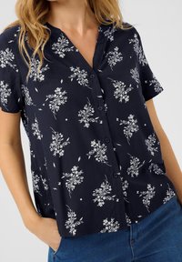 Navy short-sleeve blouse with floral print, featuring a button-up front and collar. Soft fabric with delicate white floral patterns.