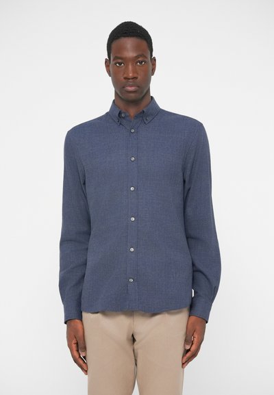 Camicia blu navy con tessuto testurizzato, caratterizzata da un colletto classico, maniche lunghe e sottili dettagli in abbottonatura. Abbinata a pantaloni beige.