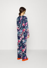 Macacão floral em azul-marinho com padrões vibrantes em rosa, roxo e verde. Mangas longas, corte solto, com sandálias laranja. Tecido suave e leve.