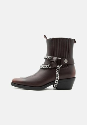 Bota de cuero marrón oscuro hasta el tobillo con tacón bajo, detalles de cadena y correa plateados, y tiradores en los laterales superiores.