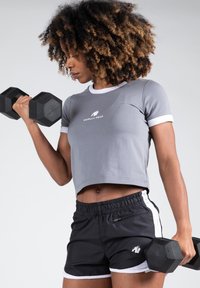 Grijs cropped sportshirt met witte rand en een logo, gecombineerd met zwarte shorts met witte zijstrepen en een klein logo, terwijl hij dumbbells vasthoudt.