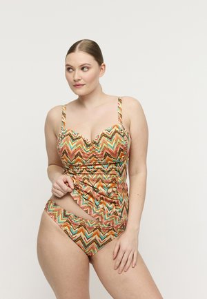 Vrouw draagt een multicolor zigzag-gepatroneerde tankini, tilt het bovenstukje licht omhoog, staand tegen een effen lichte achtergrond.