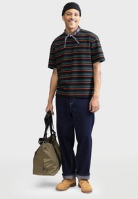 Camisa a rayas de manga corta en verde oscuro con rayas rojas y azules, combinada con unos vaqueros oscuros holgados y botas amarillas, sosteniendo un bolso en verde oliva.