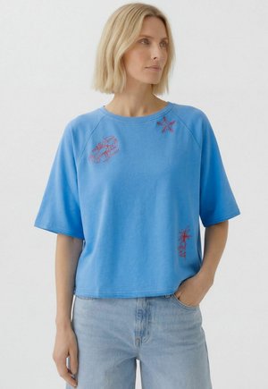 Frau trägt ein locker sitzendes blaues Kurzarmhemd mit rot besticktem Hummer, Seestern und Palme, kombiniert mit hellblauen Jeans.