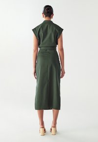 Abito verde, su misura, con maniche corte a cappuccio, cintura in vita e orlo fino al ginocchio. Tessuto liscio con una silhouette aderente. Tacchi a blocco in legno.