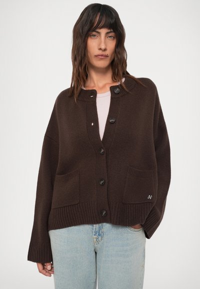 Femeie cu păr brunet, purtând un cardigan butonat maro închis cu buzunare frontale, peste o cămașă deschisă la culoare și blugi bleu deschis, pe un fundal alb.