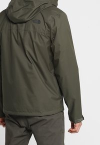 Grøn vandtæt jakke med hætte, med en logo på ryggen, justerbare manchetter og en glat, tekstureret finish.