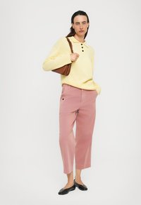 Woman wearing yellow buttoned sweater, pink wide-leg pants, black flats, and carrying a brown shoulder bag, standing against a plain white background.