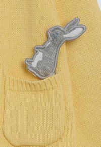 Geltonas megztinis su pilku siuvinėjimu kiškučiu, įdėtas į priekinį kišenę, minkšta tekstūra ir raštuotas apykaklė.