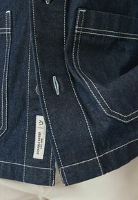 Mørkeblå denimskjorte med hvidt stikningssyning, to brystlommer og et mærke med teksten "Durable Goods" på sidesømmen.