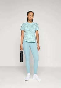 Mujer con camisa de manga corta azul claro de estampado y leggins, zapatillas blancas, sosteniendo una botella de agua negra, de pie contra un fondo liso.