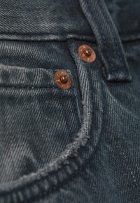 Κοντινό πλάνο σκουρόχρωμου μπλε τζιν υφάσματος με χάλκινα καρφιά και ορατή ραφή στην άκρη της τσέπης ενός τζιν.