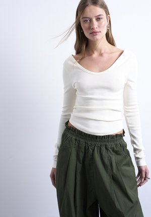 Nuori nainen, jolla on pitkät hiukset ja päällään valkoinen, olkapäät paljastava pitkähihainen paita sekä vyötäröltä korkeavyötäröiset oliivinvihreät housut, yksinkertaista taustaa vasten.